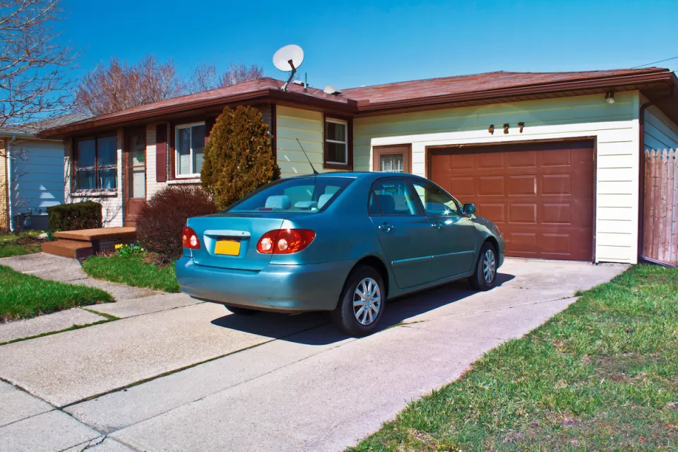 Blue small sedan in driveway of ranch-style one-story home with light green walls and dark brown garage door and trim.